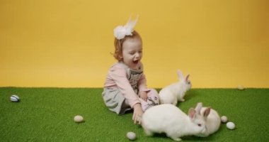 2 yaşında bir çocuk yeşil halıda tavşanlarla oynuyor ve izole edilmiş sarı arka planda renkli yumurtalar var. Mutlu Paskalyalar. Şirin tüylü tavşan, tapılası hayvan konsepti. Mutlu Paskalyalar.