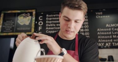 Önlüklü erkek barista çaydanlıktan bardağa su dolduruyor. Barista özel kahve dükkanında latte yapıyor. Kahve yapıyorum. Barista kahve hazırlıyor. Latte hazırlama.