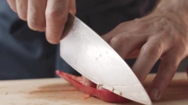 Front view of the young chef cut thin slices of a piece of red pepper with a knife on the cutting board. Front view of a young chef cutting thin slices of a piece of red pepper. Vegetables up close.
