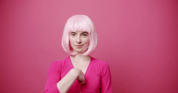 胸やカメラを指し 私はピンクの背景に笑顔でカメラを見ている間 私はあなたを愛して言うために心をジェスチャー明るい髪を持つ楽観的な若い女性の静的なショット — ストック動画