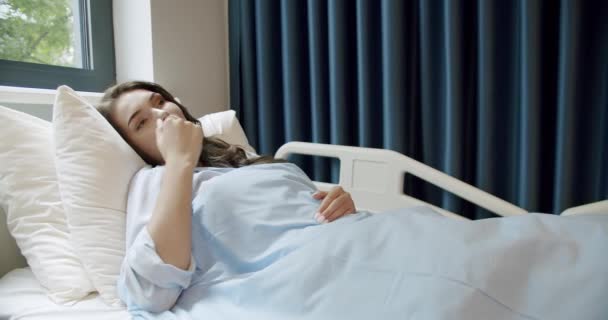 病院のベッドで女性患者 薄暗い髪の若い病気の女性のビューは 光病院病棟の窓の近くのベッドの上により快適に横たわっている 病院病棟ベッドの女性患者 — ストック動画