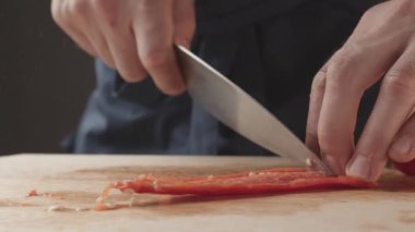 Front view of the young chef cut thin slices of a piece of red pepper with a knife on the cutting board. Front view of a young chef cutting thin slices of a piece of red pepper. Vegetables up close.