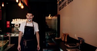 Positive young bearded male barista in white t shirt apron and reusable face mask smiling and looking at camera while standing in modern coffee house with folded arms. Confidence and lifestyle.