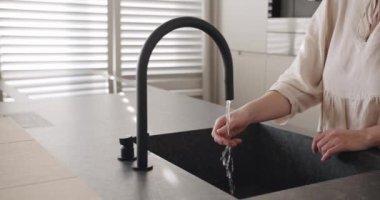 A woman turns on the tap in a modern white kitchen. Modern kitchen room in minimalist apartment, Black Kitchen sink and faucet, comfortable equipment.