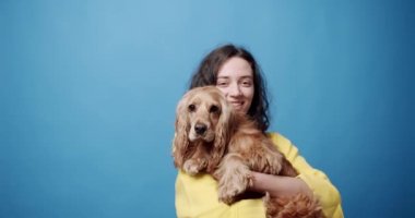 Şık giyinmiş neşeli genç esmer İngiliz Cocker Spaniel köpeğini kucaklıyor ve dişlek gülüşüyle kameraya bakıyor sonra da evcil hayvanı mavi arka plana hafifçe fırlatıyor.
