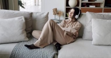 Mindful young happy woman wearing wireless headphones, listening relaxing music, resting on comfortable sofa, enjoying interesting audio book or affirmations, spending leisure indoors