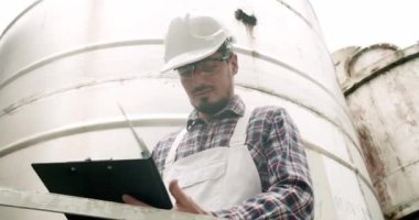 Controls the quality of production equipment in wine factory. Male wine maker checking quality of beverage distillation process indoors and write down the dates in the diary.Alcohol production concept