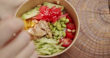 A woman hand serves Poke bowl, traditional Hawaiian raw fish salad. Poke bowl with shrimp, rice, avocado, mango, edamame beans, cucumber on a wooden background. Healthy food, keto diet concept.