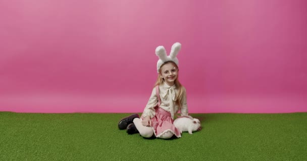 笑顔でカメラを見ているウサギの耳とメガネを持つ肯定的なブロンドの女の子と偽の芝生のピンクの背景にふわふわのウサギを撫でています ハッピーイースター ハッピーイースターの日 テキストのバナーと場所 — ストック動画
