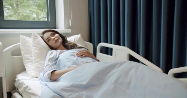 薄暗い髪の若い病気の女性のビューは 光病院病棟の窓の近くのベッドの上により快適に横たわっている 病院病棟女性患者ベッドで 病院の部屋に横になっている若い女性患者 — ストック動画