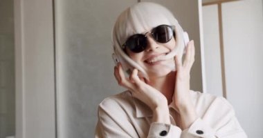 Beyaz saçlı mutlu genç bir kadın beyaz kablosuz kulaklıkla favori pop enerjik müziğini dinliyor. Mobil uygulamadan, siyah güneş gözlükleriyle. Evde keyfin yerinde. Favori bir ses parçasını dinlerken.