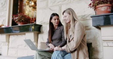 Laptop kullanan ve parmaklarına güvenen neşeli bir esmerin el kamerasıyla çekilmiş fotoğrafı. Elinde kahveyle bankta oturan ve şehir caddesinde uzaktan çalışan sarışın bir kız arkadaşın fotoğrafı.