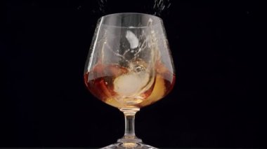 Luxury brandy. Glass with golden cognac on isolated black background with ice cube. Brandy, cognac, and whisky. Slow motion. Luxury Drink Whisky with smoke. The ice falls into the whiskey glass.