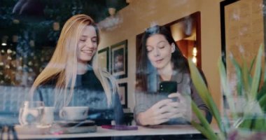 Kafede muhabbet eden mutlu kız arkadaşlar. Pencerenin arkasındaki kafeterya masasında oturmuş iyimser sarışın kız arkadaşına el kamerasıyla el hareketleriyle hikâyeler anlatan genç bir kadın.