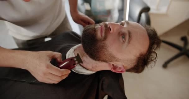 一个英俊的男人用电动理发机改头换面 近距离与修剪工作 年轻的嬉皮士白种人在现代理发店修整胡子和胡子 男人的发型 — 图库视频影像