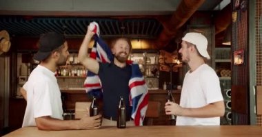 Büyük Britanya bayrağı taşıyan erkekler bira içip spor maçı izliyorlar. Taraftarlar bir barda arkadaşlarıyla futbol izliyorlar. Sadık futbol taraftarları takımlarını destekliyor..