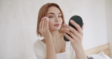 Genç esmer kadın küçük yuvarlak aynaya bakar ve yüzündeki makyajı pamukla temizler. Cilt bakımı. Cilt bakımı ve temizlik prosedürü. Losyonla cilt nemlendirici.