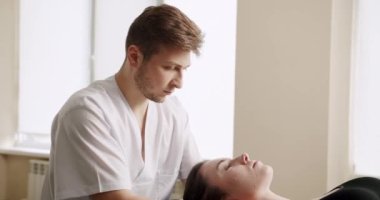 Masaj terapisti bayan müşteriyi tedavi ediyor. Güneşli klinik ofisinde masaj sırasında erkek osteopatın, kadının boyun kaslarını yoğurduğu el kamerası görüntüsünü yakınlaştır..
