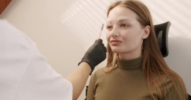 Usta beyaz işaretleri özel bir fırçayla kaşların şeklini düzeltmek için uygular. Mikroplu patinaj yapmadan önce güzellik salonundaki kanepede oturan genç bir kızın güzel yüzü, kalıcı makyaj kaşlı dövme..