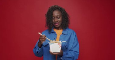 Afro-Amerikalı kadın kameraya bakıyor ve gülümsüyor, izole edilmiş kırmızı arka planda erişte, deniz ürünü ve sebze yemeye hazırlanıyor. Asya sokak yemekleri.