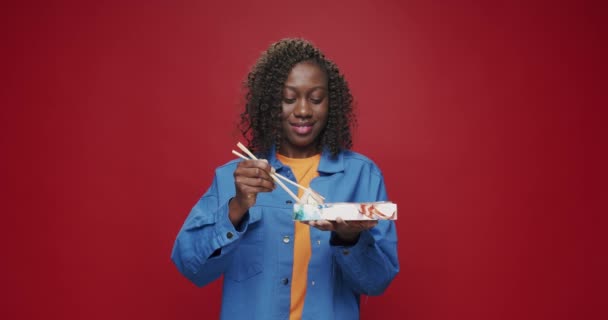 アフリカ系アメリカ人の女性はカメラを見て笑顔で 孤立した赤を背景に箸で寿司を食べる準備をしています 寿司ロール — ストック動画