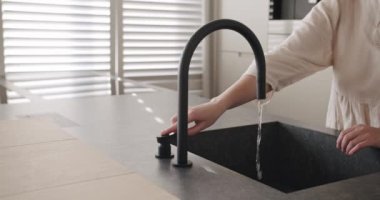 A woman turns on the tap in a modern white kitchen. Modern kitchen room in minimalist apartment, Black Kitchen sink and faucet, comfortable equipment.