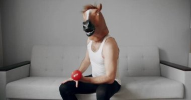 Komik Sağlık ve Spor Konsepti. Esprili esnemeler. At maskeli komik bir adam evdeki beyaz koltukta ağırlık çalışması için egzersiz yapıyor. Evde komik bir spor salonu var. Komik ev egzersizi.