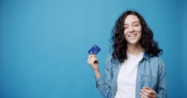 クレジットカードで幸せな若いブルネット 若い女性銀行クライアントの静的なショットでカメラを見て笑顔とシャーククレジットカードで青の背景 — ストック動画