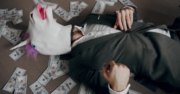 スーツでユニコーンマスクを持つ面白い男は 彼の財政的な成功を楽しんで 床の上に座ってお金を数える ハンサムな幸せなビジネスマンとともにユニコーンマスク円を散布 — ストック動画