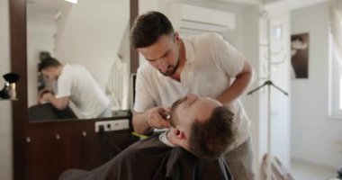 Yakışıklı adam elektrikli budayıcı ile yeni bir saç stili alıyor. Makyaj işiyle kapat. Modern berber dükkanında sakal ve bıyık bakımında genç hippi beyaz adam. Erkek saç stili. 