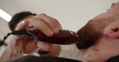 Yakışıklı adam elektrikli budayıcı ile yeni bir saç stili alıyor. Makyaj işiyle kapat. Modern berber dükkanında sakal ve bıyık bakımında genç hippi beyaz adam. Erkek saç stili. 