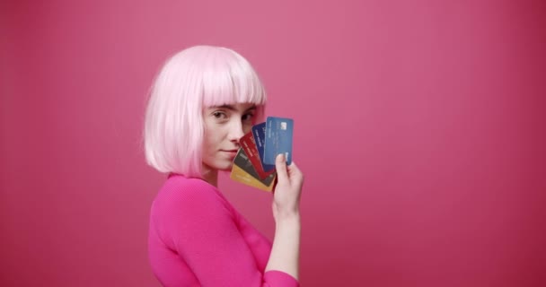 クレジットカードを見せる若い女性 若いです女性銀行クライアントの静的なショットとともに明るい髪デモンストレーション束のカラフルなクレジットカードに対してピンクの背景 — ストック動画