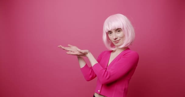时髦的年轻女性指指点点 身着时髦服装 头戴无线耳机 头戴明亮头发 指着粉红背景微笑的友善年轻女子的静态照片 — 图库视频影像