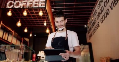 Beyaz tişörtlü, yoğun sakallı erkek barista siyah önlüklü ve tablet kullanan şeffaf yüz maskeli modern kahve dükkanında çalışırken kayıt yaptırıyor.