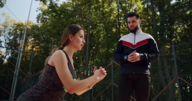 Antrenör erkek, formda kadın egzersizleri yapıyor. Kişisel antrenörle açık hava eğitimi. Genç kız ve erkek egzersiz yapıyor. Sportif erkek ve kadın kaslarını güçlendirmek için tek ayak üzerinde hamle yapıyorlar.