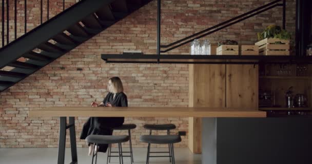 这个女人坐在宽敞的现代木制厨房的椅子上 房间里有墙砖和黑色楼梯 房地产 新家的简约设计 真正的设计内部 新古典主义风格 舒适的内部 — 图库视频影像