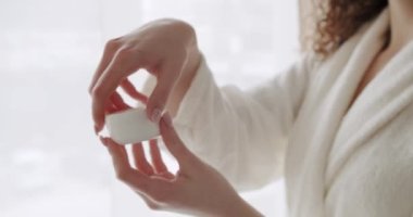 Ellerinde bornoz olan genç bir kadın yüz kremiyle bir şişe açıyor ve evde kokuyor. Beyaz krema kavanozunu açtım. Tedavi gören bir kadın vücut bakımını, sağlığını ve sağlığını nemlendiriyor.