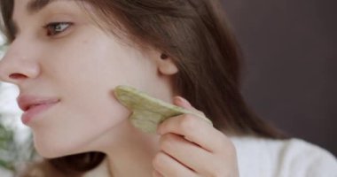 Yeşil bir Gua Sha taş kazıyıcısıyla yüz masajı yapan genç bir kadının portresi. Evde güzellik tedavisi. Kadın anti-reşit yüz masajı için gua sha yeşim kazıyıcı kullanıyor..
