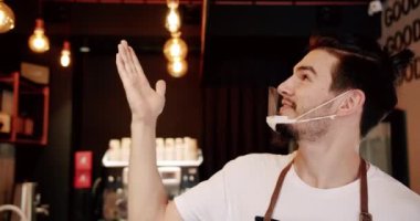 Barda duran bir işyeri sahibi. Olumlu genç barista gülümseyen ve kameraya bakarken modern kafede neler olduğunu gösteriyor. Kahve dükkanında çalışırken gülümseyen genç adam..
