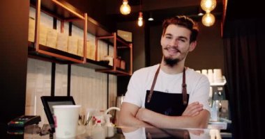 Beyaz tişörtlü, önlüklü, sakallı, modern kafe tezgahında aperatif servisi yaparken gülümseyen ve kameraya bakan genç bir barista.