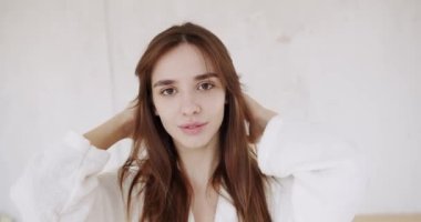 Sağlıklı kuru cilt bakımı, cilt bakımı, cilt bakımı, beyaz bornozlu genç bir kadın nazikçe yüzüne gözünün altından boynuna dokunuyor ve kameraya bakıyor.