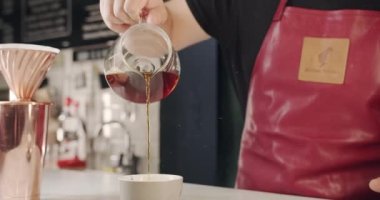 Tezgahta kahve barmeninin bardağa kahve doldurması. Kırmızı önlüklü meçhul erkek barista modern açık kafede dururken yeni hazırlanmış filtre kahveyi fincana dolduruyor..