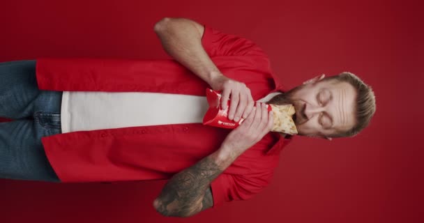 Unshaven白人男性の垂直ビデオは 孤立した赤の背景に喜びを持って肉ケバブを食べ 彼の口でシャワルマをかむ 男は気軽にファーストフードからジャンクフードを食べる 不健康な食べ物 ジャンクフード — ストック動画