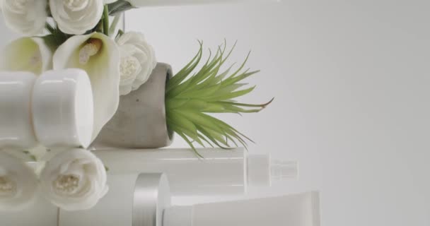スタジオの白い背景に化粧品のための美しさの花を持つ白いボトルとチューブ 天然成分 スクラブ 強壮剤 ボディケアに基づく化粧品油 ストーリーの垂直ビデオ — ストック動画