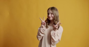 12 yıllık bej elbiseli sarışın genç kız mutlu ve gülümsüyor. Stüdyodaki sarı arka planda zafer işareti gösteriyor. Genç kız gülümsüyor ve iki parmağını gösteriyor, zafer işareti..