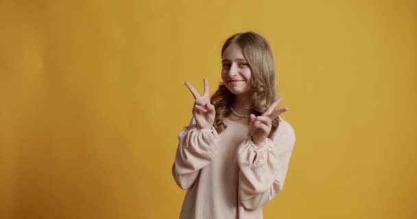ブロンドのティーンエイジャーの女の子12ベージュのドレスで年彼女はスタジオで黄色の背景に幸せと笑顔と勝利の兆候を示しています 10代の少女は笑顔で2本の指を見せ — ストック動画