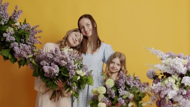 年轻的母亲抱着两个孩子 在一个黄色的背景上看着镜头 在工作室里放着自然的花朵 快乐的家庭支持 结合或照顾快乐的妈妈拥抱 爱的家庭 — 图库视频影像