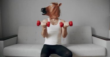 Esprili esnemeler. Komik ev egzersizi. At maskeli komik bir adam evdeki beyaz koltukta ağırlık çalışması için egzersiz yapıyor. Evde komik bir spor salonu var. Komik Sağlık ve Spor Konsepti.