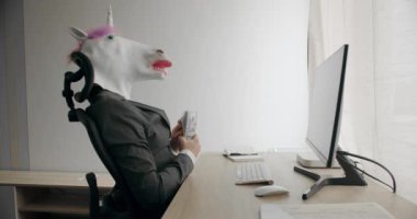 Takım elbiseli, tek boynuzlu at maskeli komik adam mali başarısının tadını çıkarıyor ve işyerinde para sayıyor. Tek boynuzlu at maskeli adam para atıyor. Para yağmuru, düşen dolarlar. Yavaş çekim.