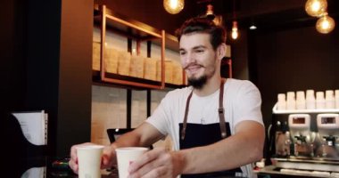 Düşük açılı, beyaz tişörtlü, önlüklü, sakallı bir erkek barista modern kafede kahve servisi yaparken gülümseyip kameraya bakıyor.